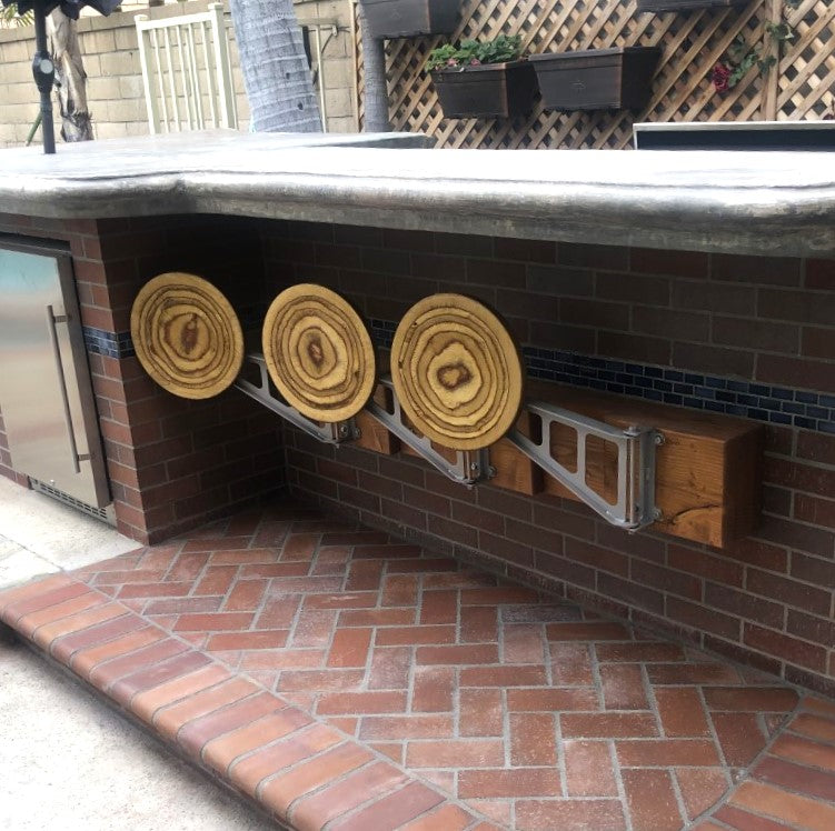 A set of three metal swing-away bar stools with wooden seats, mounted on a brick wall. The stools are in a retracted position against the wall.