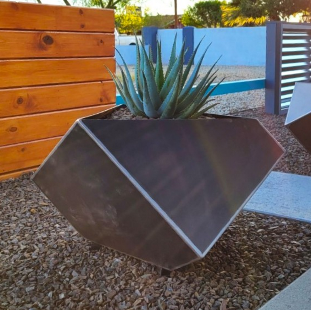 A geometric-shaped metal planter with a dark finish, placed on a gravel surface, containing a single plant.