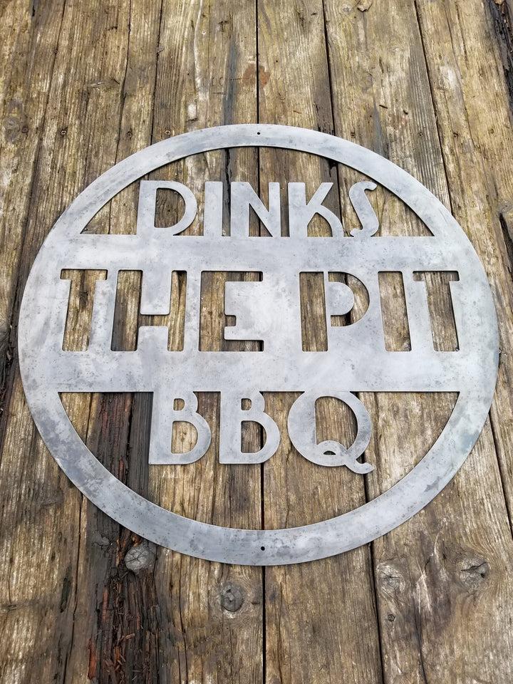This is a round art deco. sign that has three lines of text. The sign reads, " Dinks the Pit BBQ"
