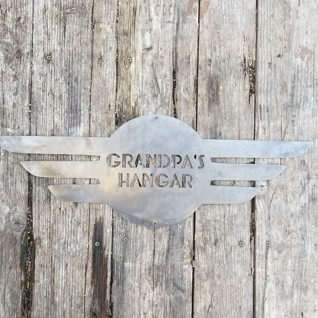 This is a round metal sign with wings. It looks like a pilot's pedant. the sign reads, "Grandpa's Hangar"