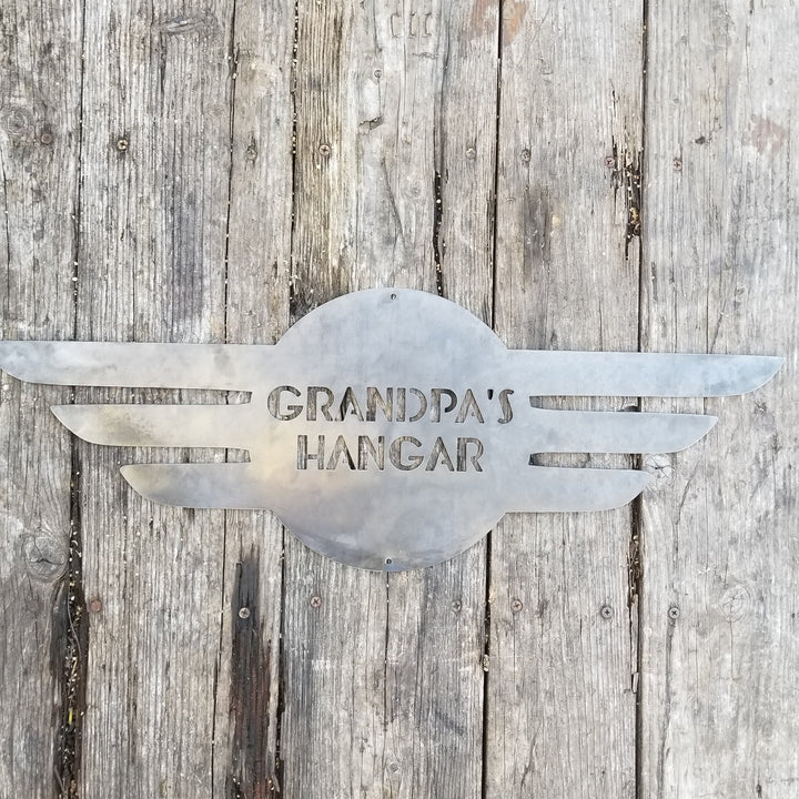 This is a round metal sign with wings. It looks like a pilot's pedant. the sign reads, "Grandpa's Hangar"