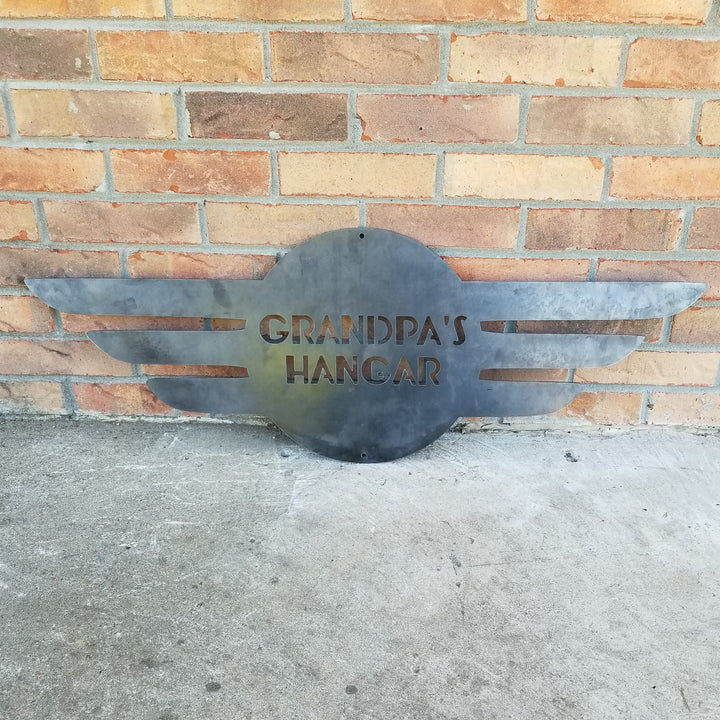 This is a round metal sign with wings. It looks like a pilot's pedant. the sign reads, "Grandpa's Hangar"