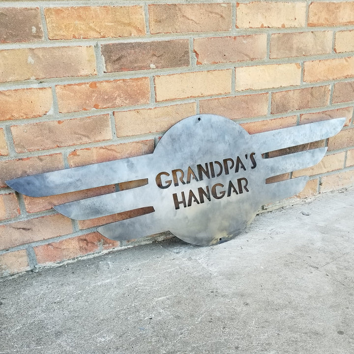 This is a round metal sign with wings. It looks like a pilot's pedant. the sign reads, "Grandpa's Hangar"