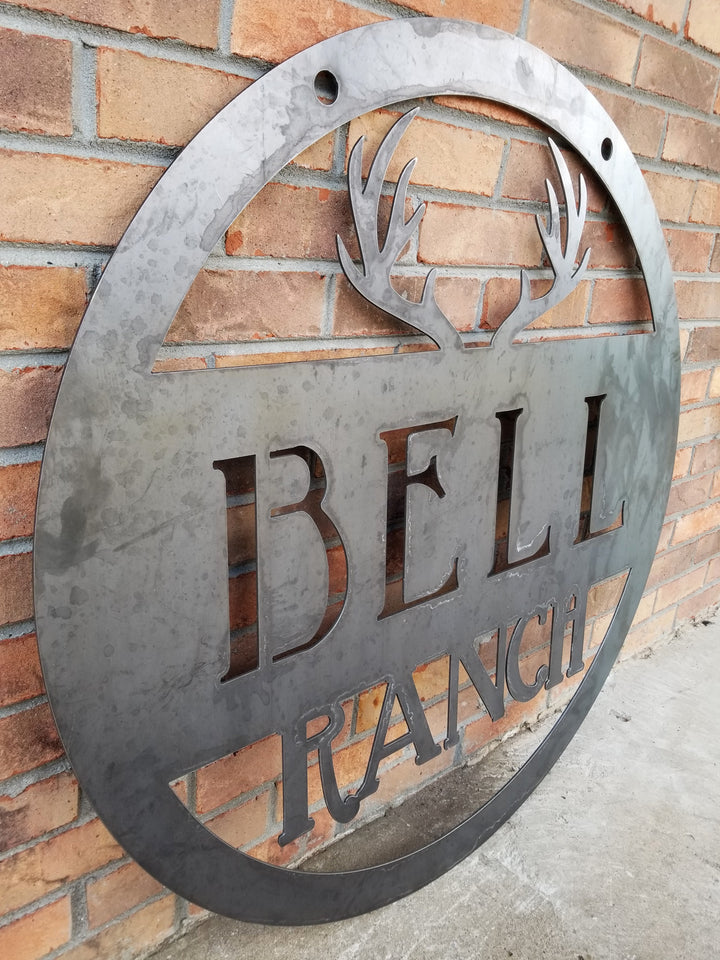 Round metal sign with an image of antlers at the top and two lines of text which read, "Bell Ranch"