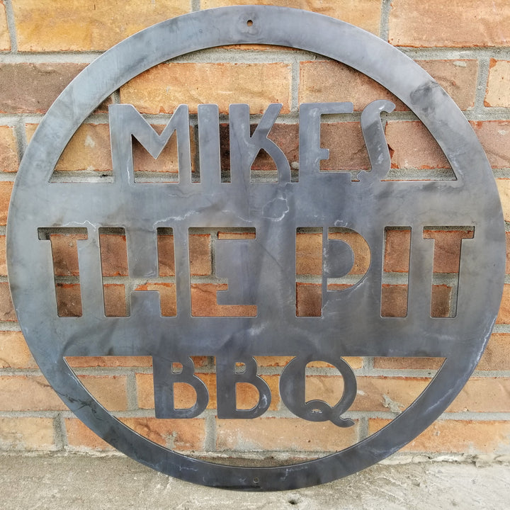 This is a round art deco. sign that has three lines of text. The sign reads, " Mikes The Pit BBQ"