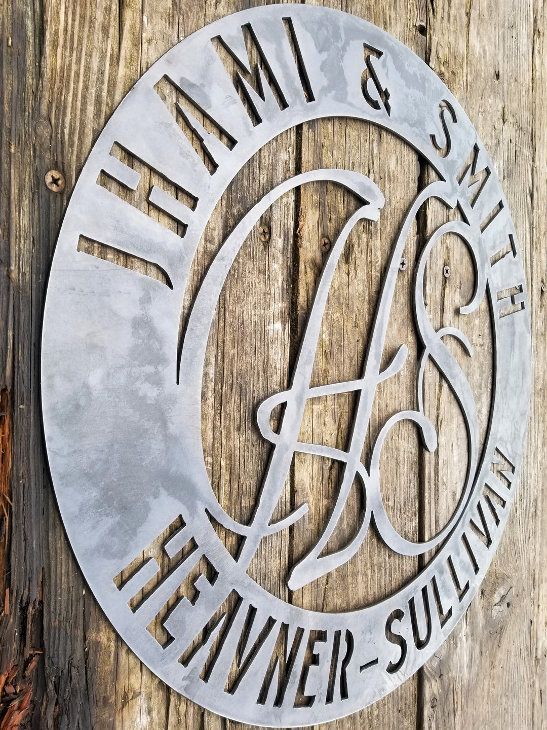 Round raw steel monogram with two lines of text and two letters in the center. The sign reads, " Jhami & Smith, Heavner-Sullivan"