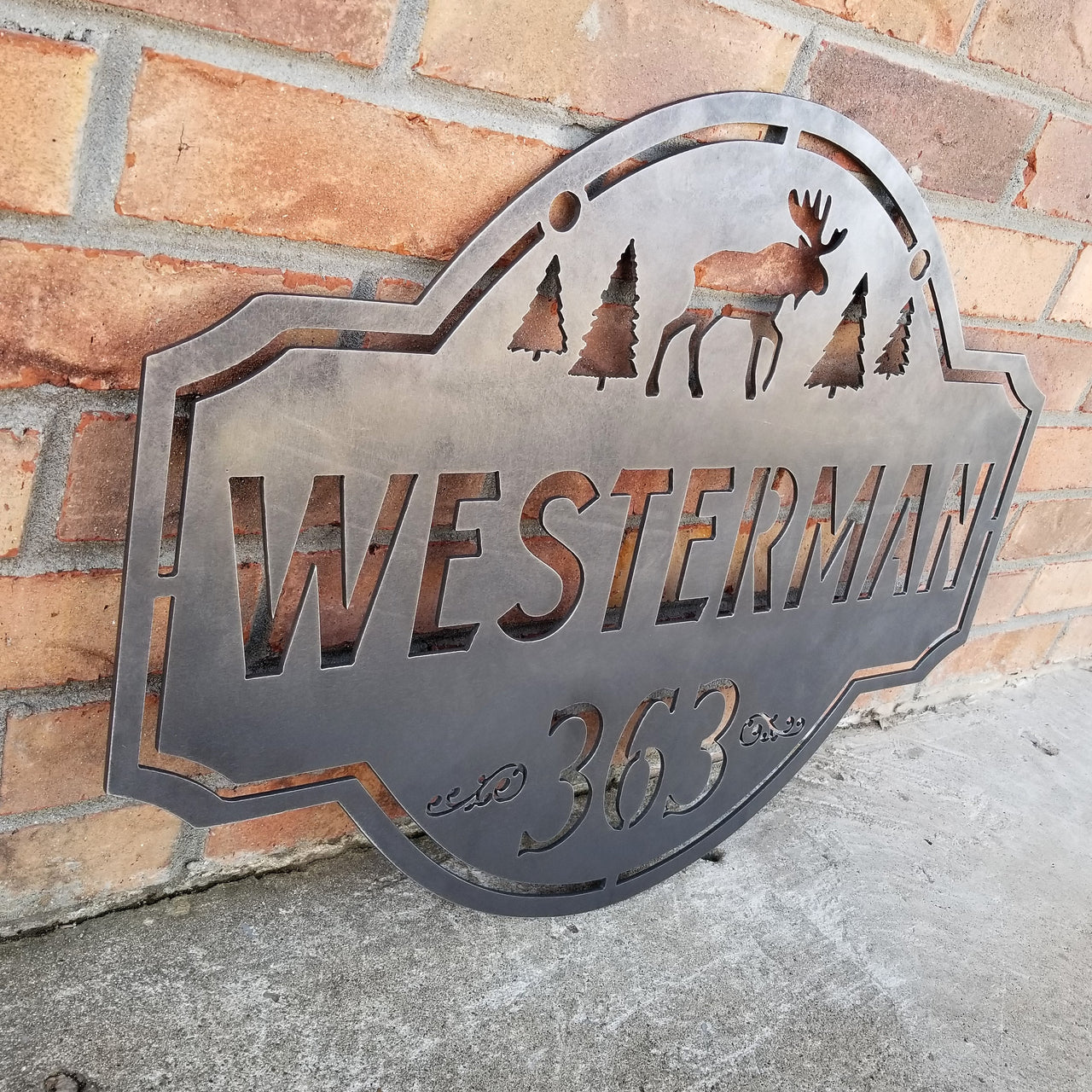 This is a personalized metal address sign that hangs from a hanging bracket mounted to a wooden post.  The sign features a forest scene with a moose in the center. The sign reads, "Westerman 363".