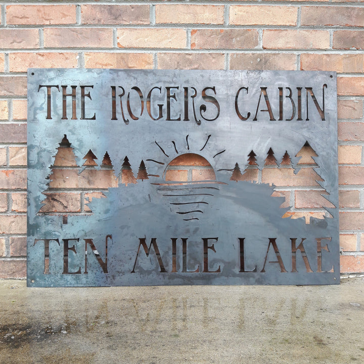 This metal sign has a tree line with a setting sun and reads, "The Rogers Cabin Ten Mile Lake"