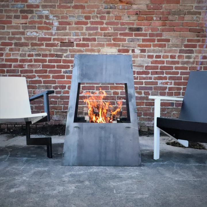 A gray steel outdoor wood burning fire pit with visible flames and wood, placed on a concrete surface with a brick wall in the background and a black modern chair to the side.