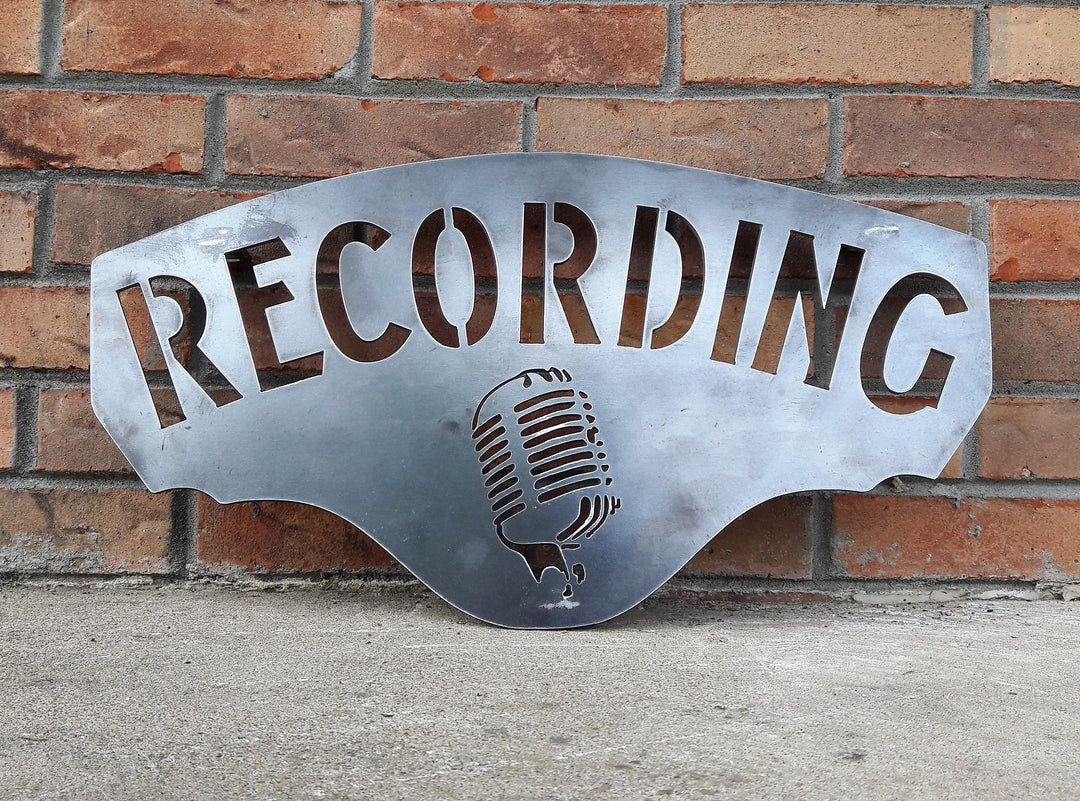 This metal sign has hidden mounts welded to the back which stands the sign 1" off the wall.  There is an image of a microphone on the sign and a line of text. The sign reads, "RECORDING".