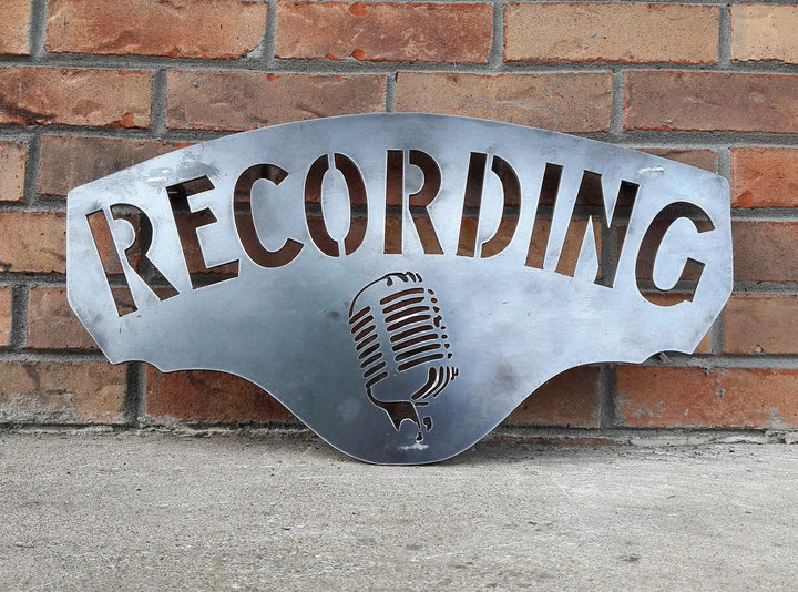 This metal sign has hidden mounts welded to the back which stands the sign 1" off the wall.  There is an image of a microphone on the sign and a line of text. The sign reads, "RECORDING".