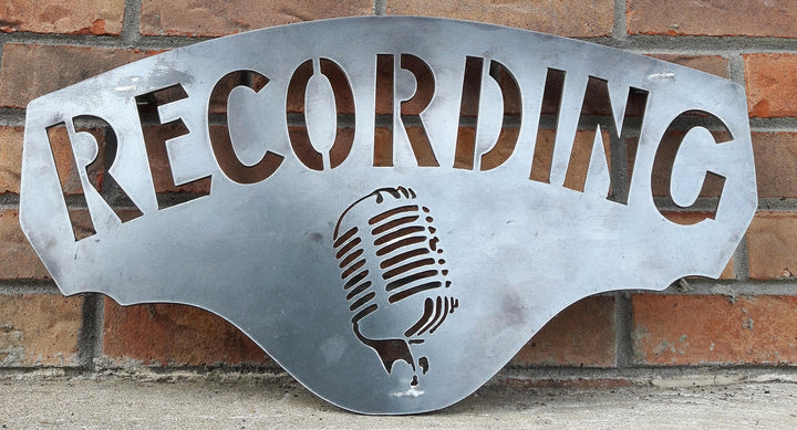 This metal sign has hidden mounts welded to the back which stands the sign 1" off the wall.  There is an image of a microphone on the sign and a line of text. The sign reads, "RECORDING".