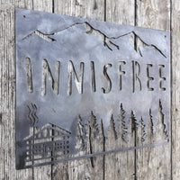 Thumbnail for This sign is displays a mountain range in the background with a forest and cabin. The sign reads, 