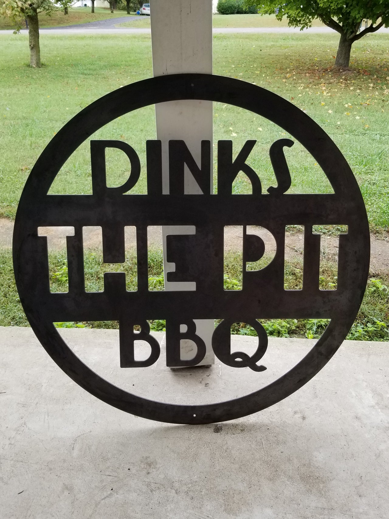 This is a round art deco. sign that has three lines of text. The sign reads, " Dinks the Pit BBQ"