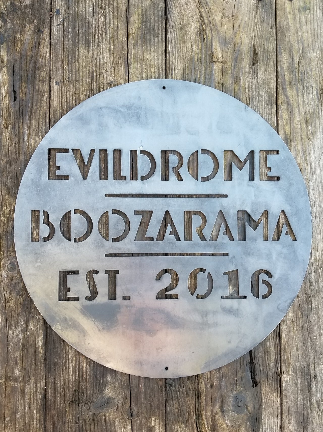 This is a round art deco sign that has three lines of text with a straight line seperating them. The sign reads, "Evildrome Boozarama Est. 2016"