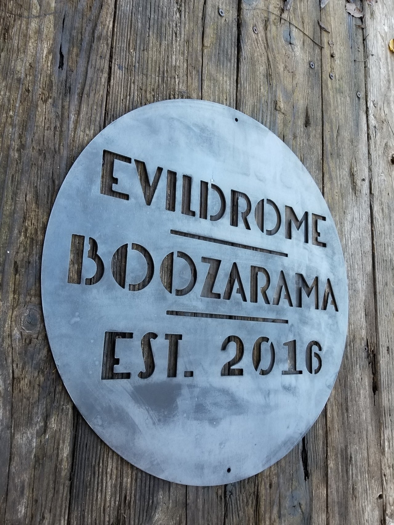 This is a round art deco sign that has three lines of text with a straight line seperating them. The sign reads, "Evildrome Boozarama Est. 2016"