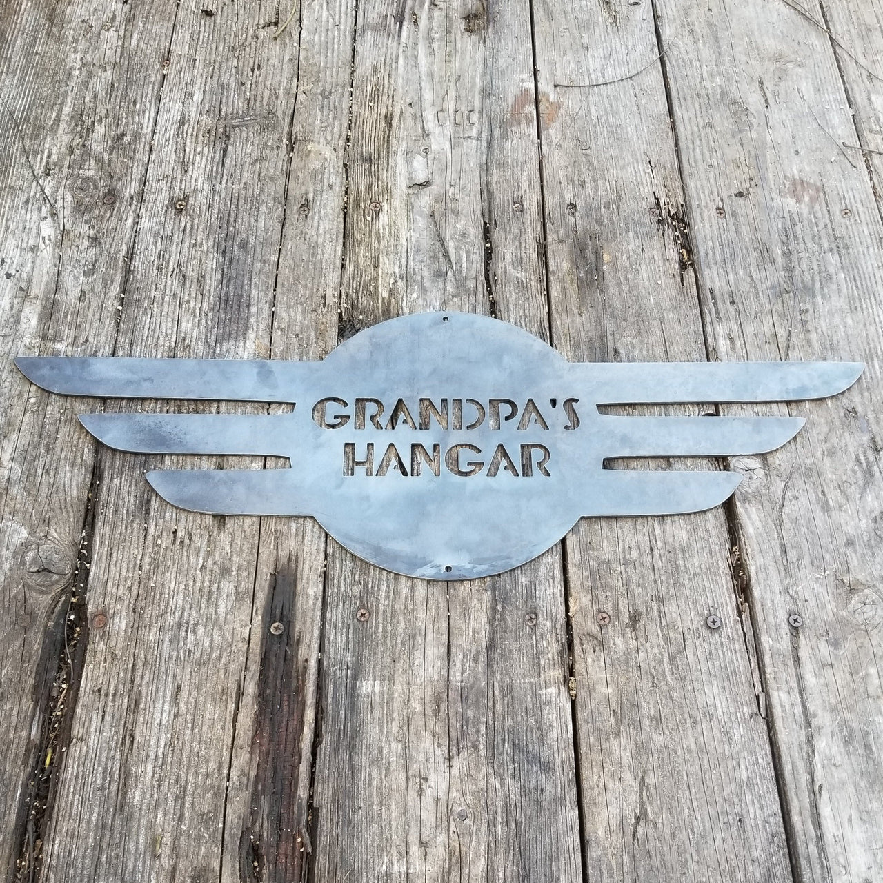 This is a round metal sign with wings. It looks like a pilot's pedant. the sign reads, "Grandpa's Hangar"