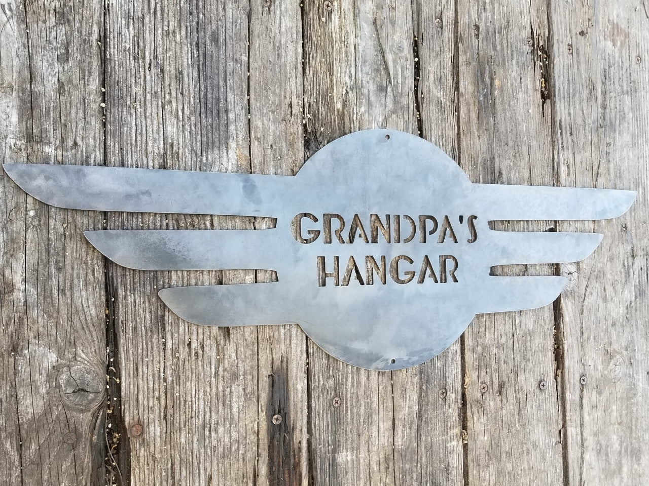 This is a round metal sign with wings. It looks like a pilot's pedant. the sign reads, "Grandpa's Hangar"