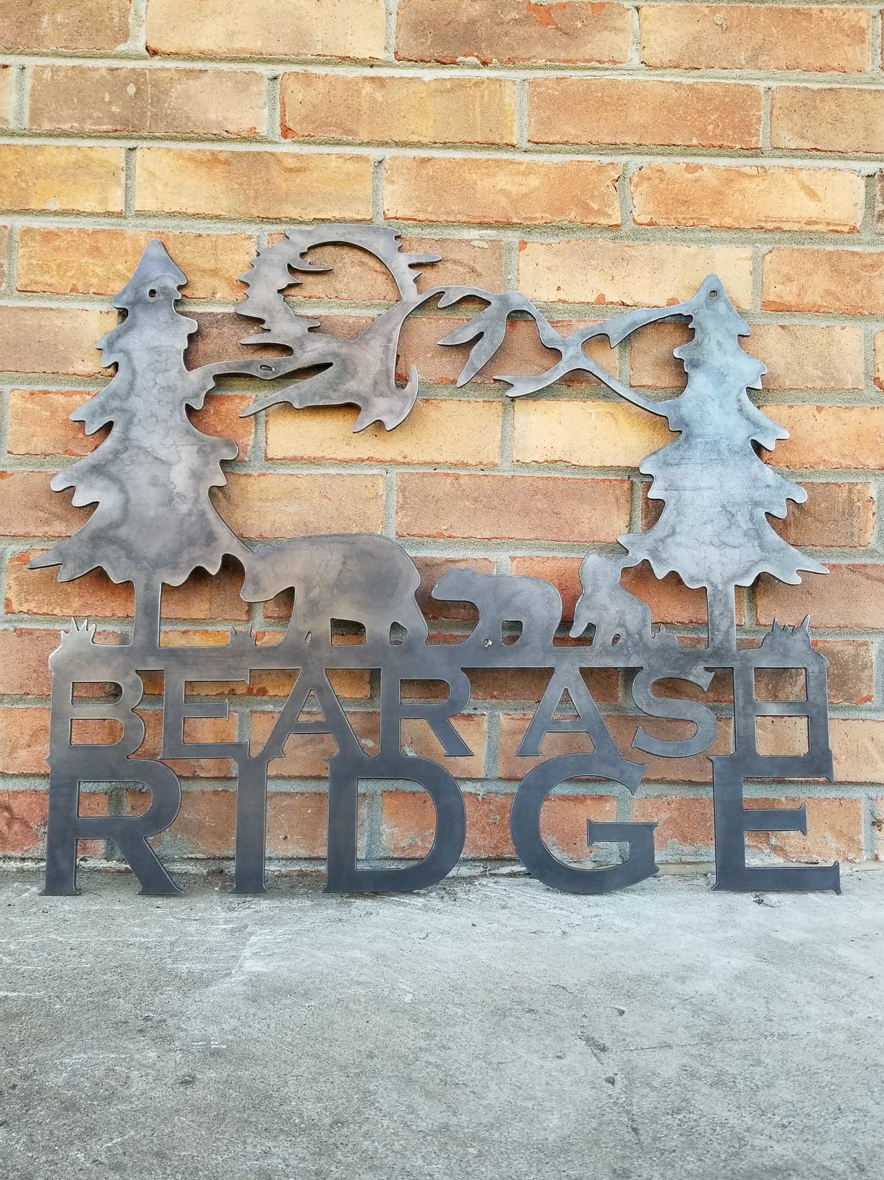 A tree scene with a family of bears and the sun in the background. Underneath the scene the metal sign reads, "Bear Ash Ridge"
