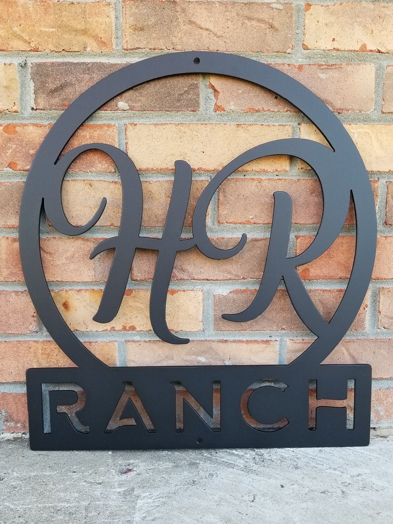 Round metal monogram with two letters at the top and a line of text at the bottom. The sign reads, " HR Ranch". The sign is powder coated black.