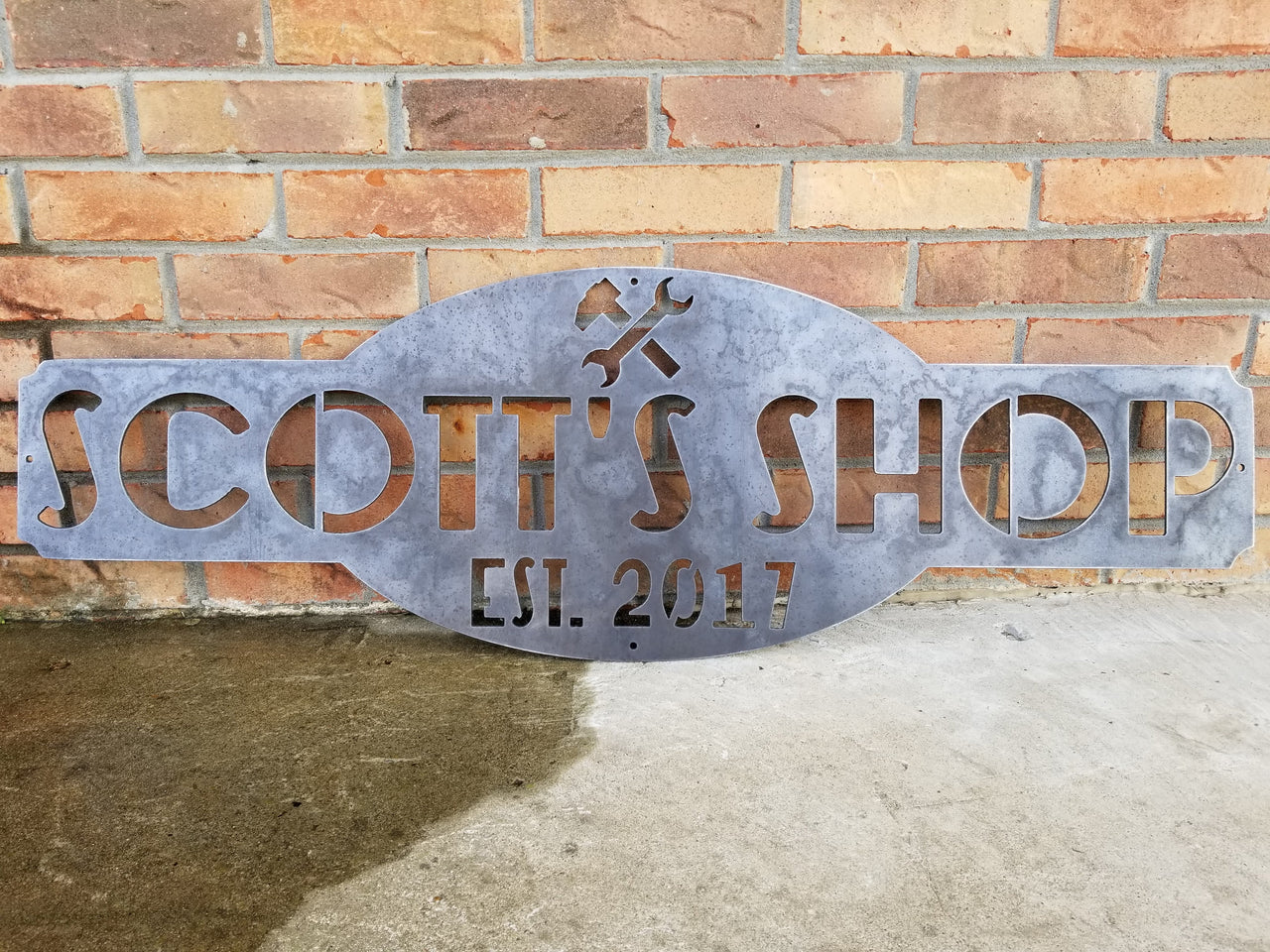 This is a raw steel, custom metal sign.  At the top of the sign is an image of tools. There is two lines of text on this sign which reads, "Scott's Shop, Est. 2013"