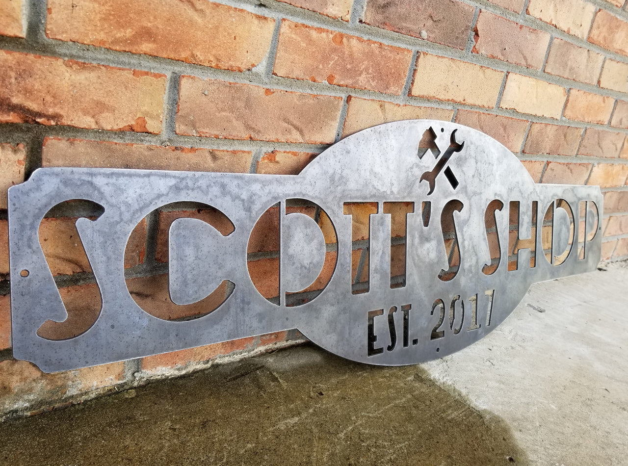This is a raw steel, custom metal sign.  At the top of the sign is an image of tools. There is two lines of text on this sign which reads, "Scott's Shop, Est. 2013"