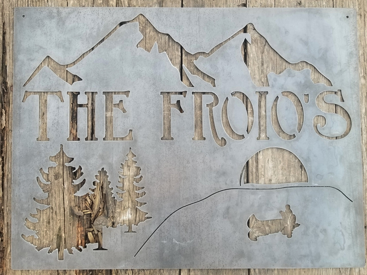 This is rectangular sign that displays a mountain range over a lake that is surrounded by trees. In the center of the lake is a boat with the figure of a man fishing who cast a shadow by the light of a sunset. Under the mountain range is a line of text that reads, "The Froio's"