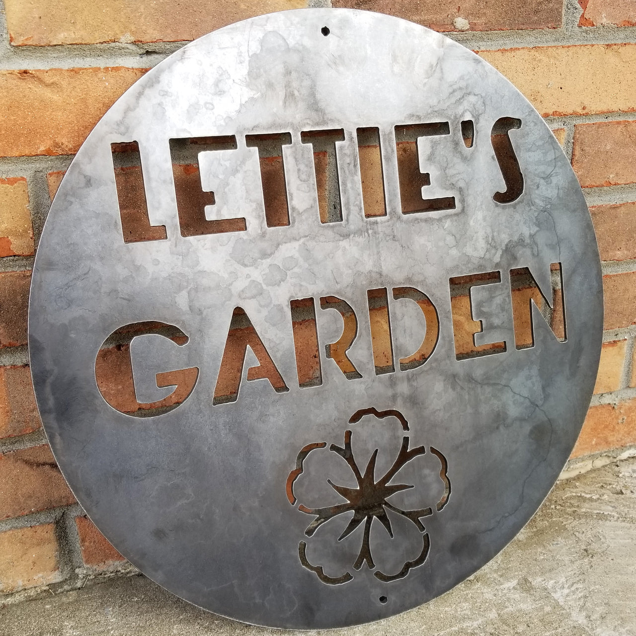 This is a round sign with an image of a flower below two lines of text which read, "Lettie's Garden".