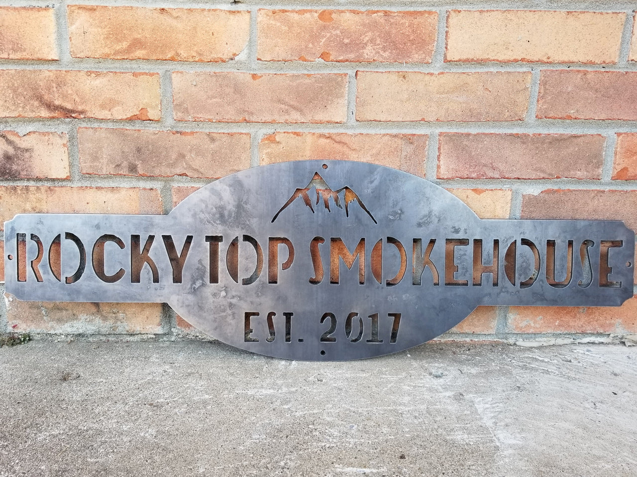 This is a raw steel, custom metal sign.  At the top of the sign is an image of mountains. There is two lines of text on this sign which reads, "Rockytop Smokehouse, Est. 2017"