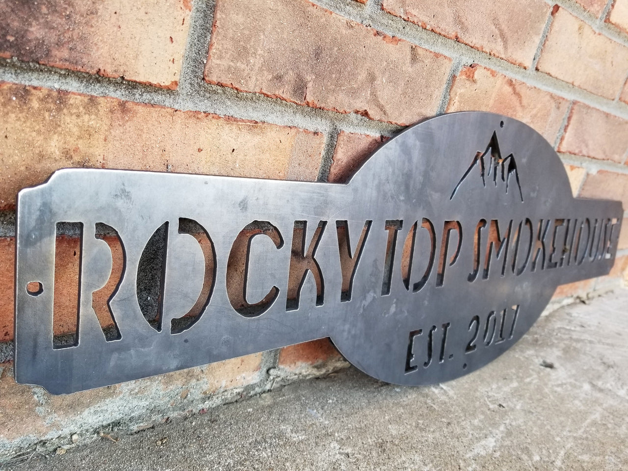 This is a raw steel, custom metal sign.  At the top of the sign is an image of mountains. There is two lines of text on this sign which reads, "Rockytop Smokehouse, Est. 2017"