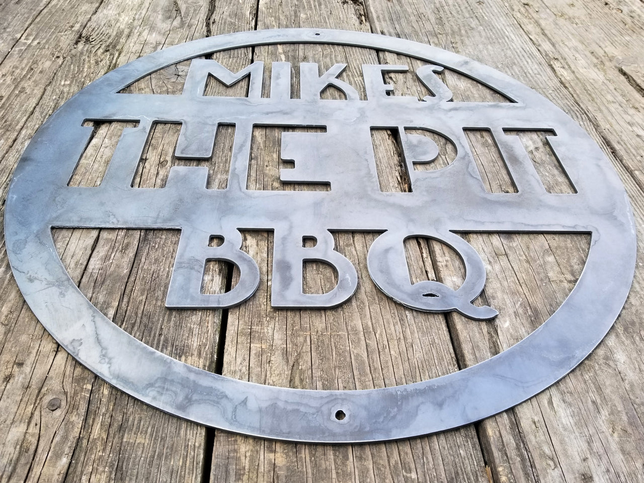 This is a round art deco. sign that has three lines of text. The sign reads, " Mikes The Pit BBQ"