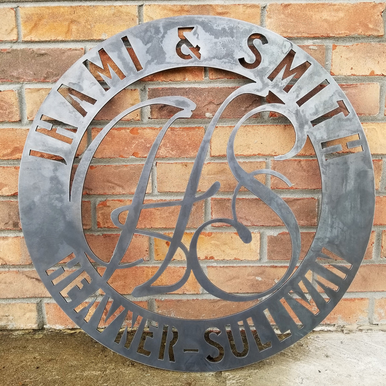 Round raw steel monogram with two lines of text and two letters in the center. The sign reads, " Jhami & Smith, Heavner-Sullivan"