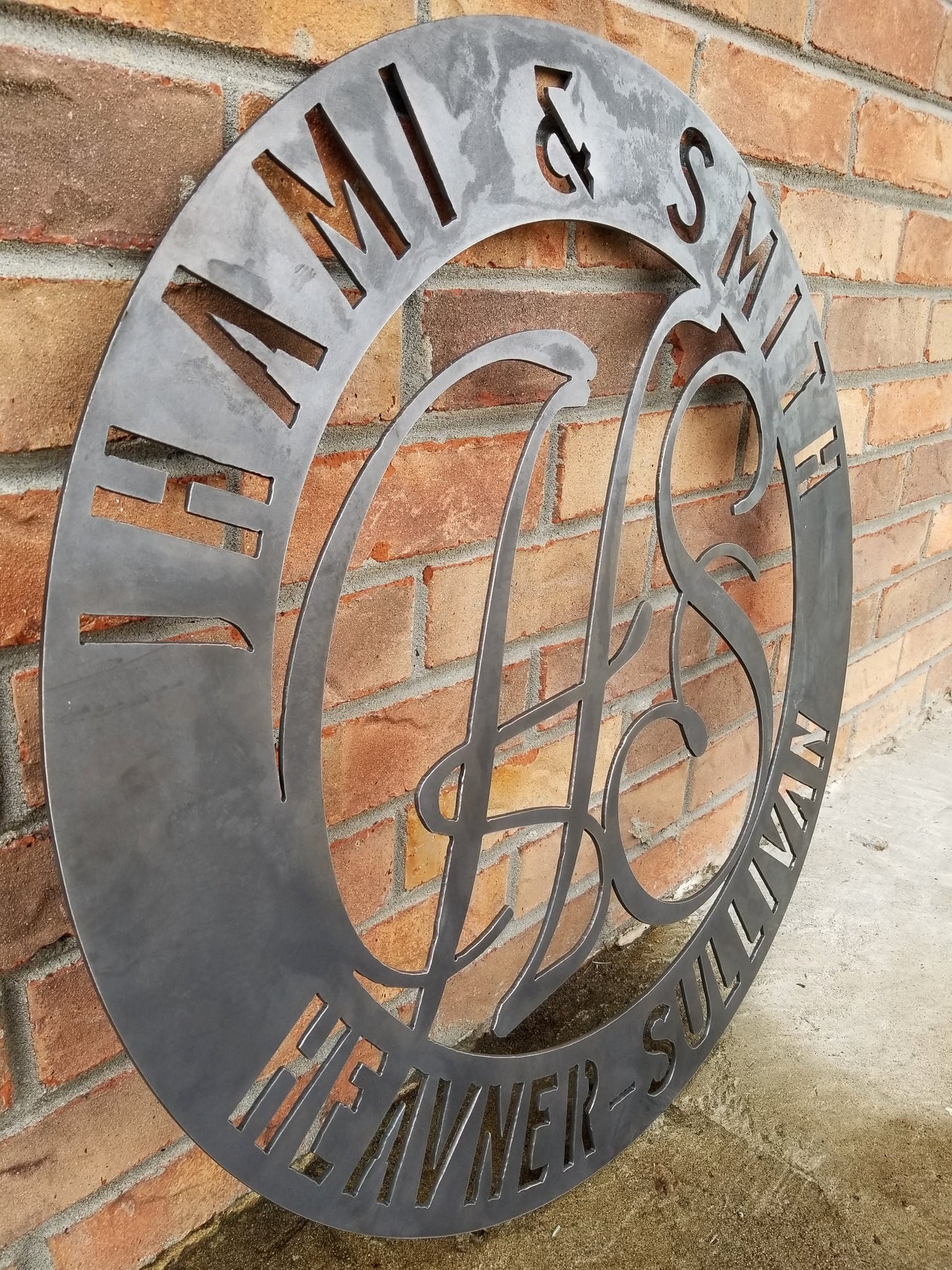 Round raw steel monogram with two lines of text and two letters in the center. The sign reads, " Jhami & Smith, Heavner-Sullivan"