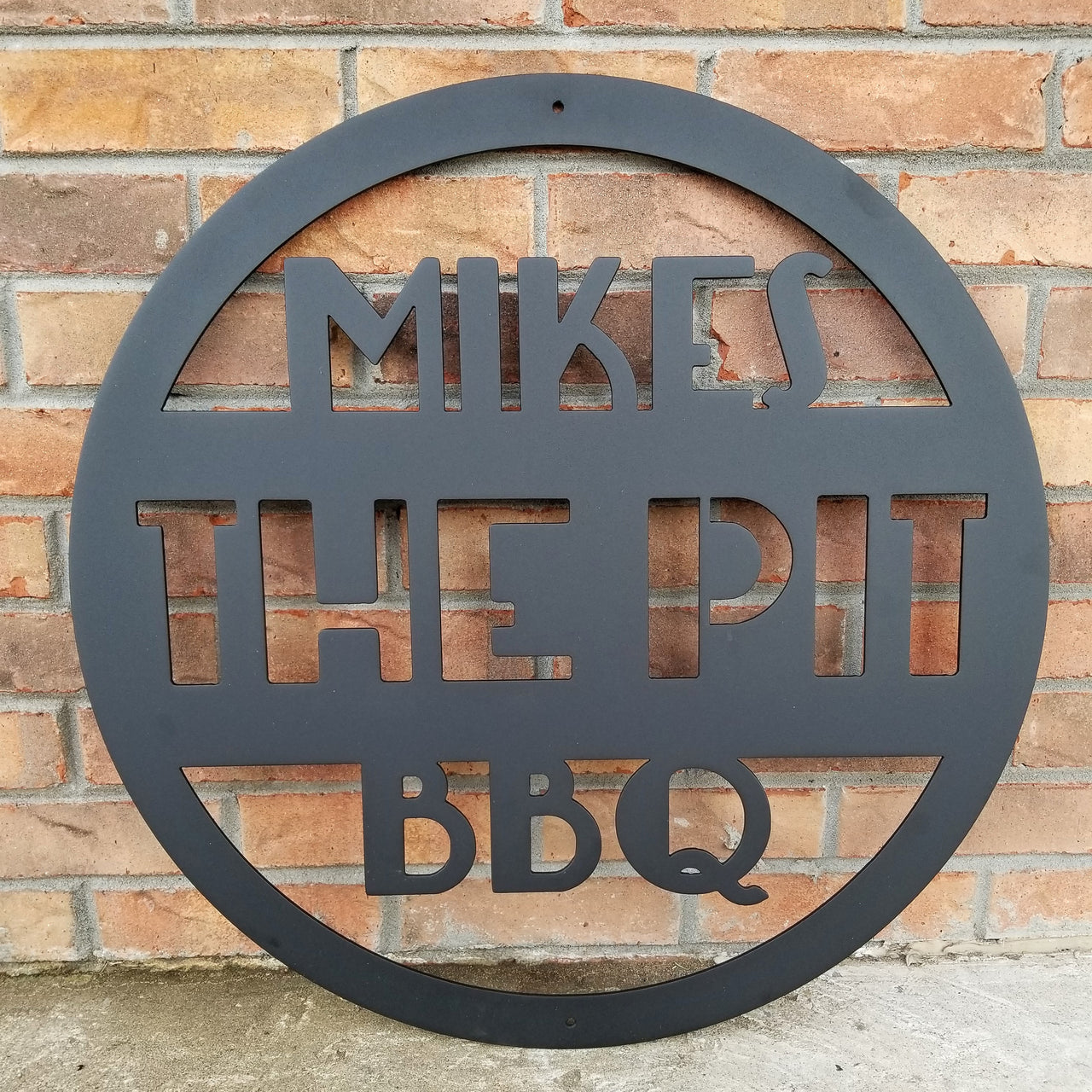 This is a round art deco. sign that has three lines of text. The sign reads, " Mikes The Pit BBQ"