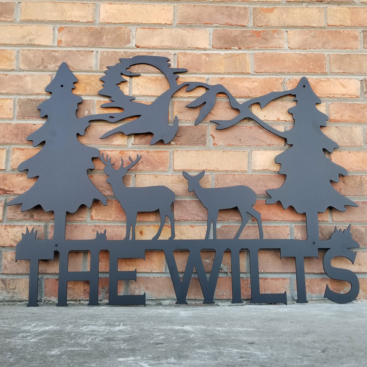 A tree scene with a family of deer and the sun in the background. Underneath the scene the metal sign reads, "The Wilts"