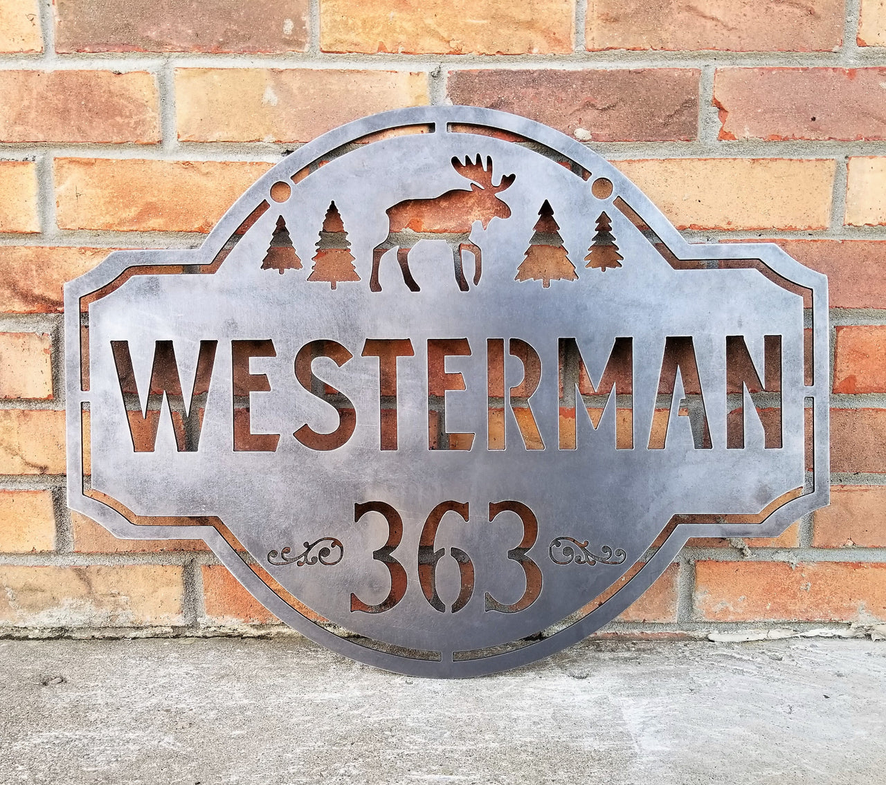 This is a personalized metal address sign that hangs from a hanging bracket mounted to a wooden post.  The sign features a forest scene with a moose in the center. The sign reads, "Westerman 363".
