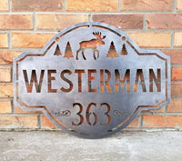 Thumbnail for This is a personalized metal address sign that hangs from a hanging bracket mounted to a wooden post.  The sign features a forest scene with a moose in the center. The sign reads, 