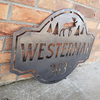 Thumbnail for This is a personalized metal address sign that hangs from a hanging bracket mounted to a wooden post.  The sign features a forest scene with a moose in the center. The sign reads, 