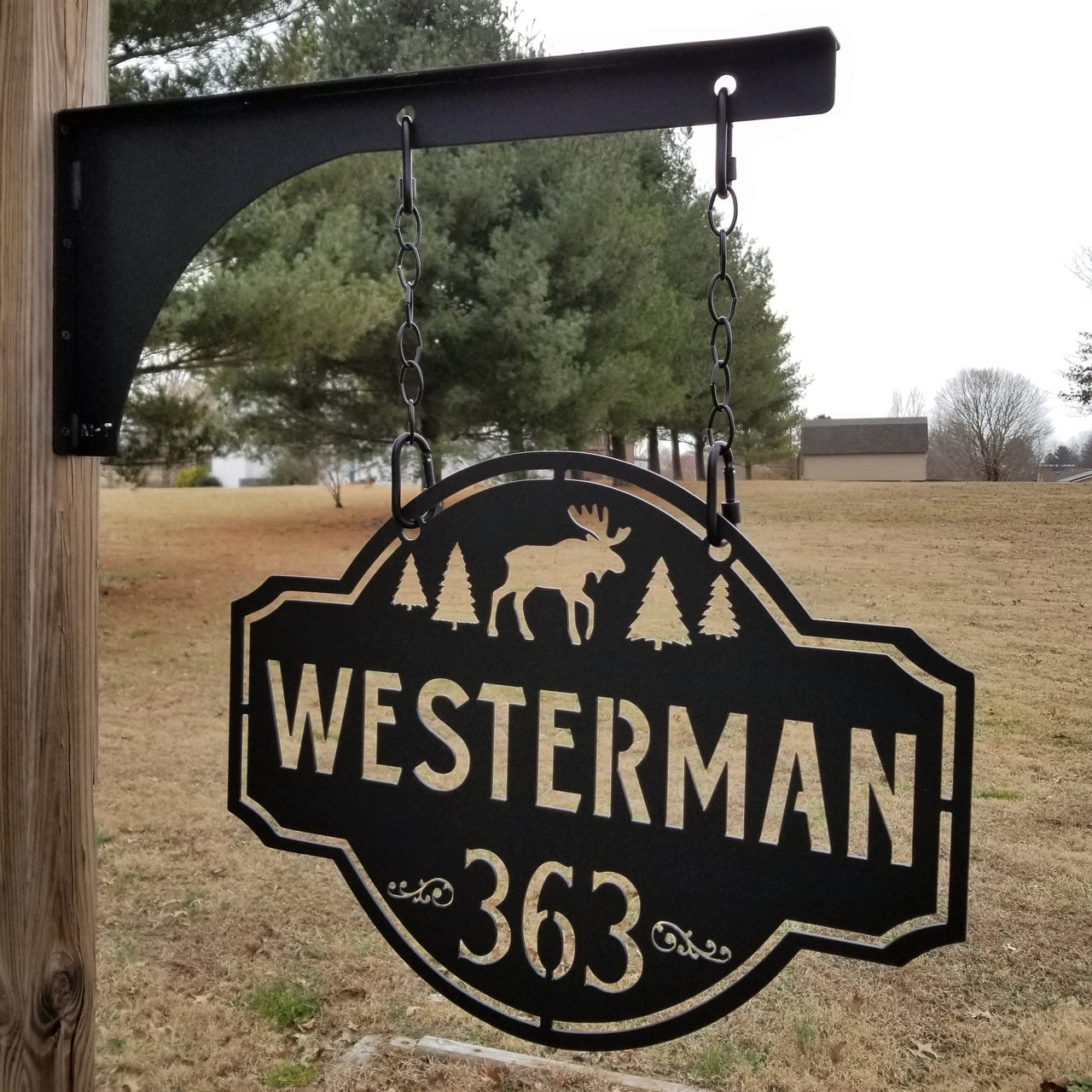 This is a personalized metal address sign that hangs from a hanging bracket mounted to a wooden post.  The sign is powder coated black and features a forest scene with a moose in the center. The sign reads, "Westerman 363".