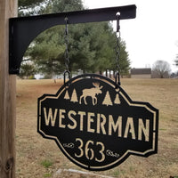 Thumbnail for This is a personalized metal address sign that hangs from a hanging bracket mounted to a wooden post.  The sign is powder coated black and features a forest scene with a moose in the center. The sign reads, 