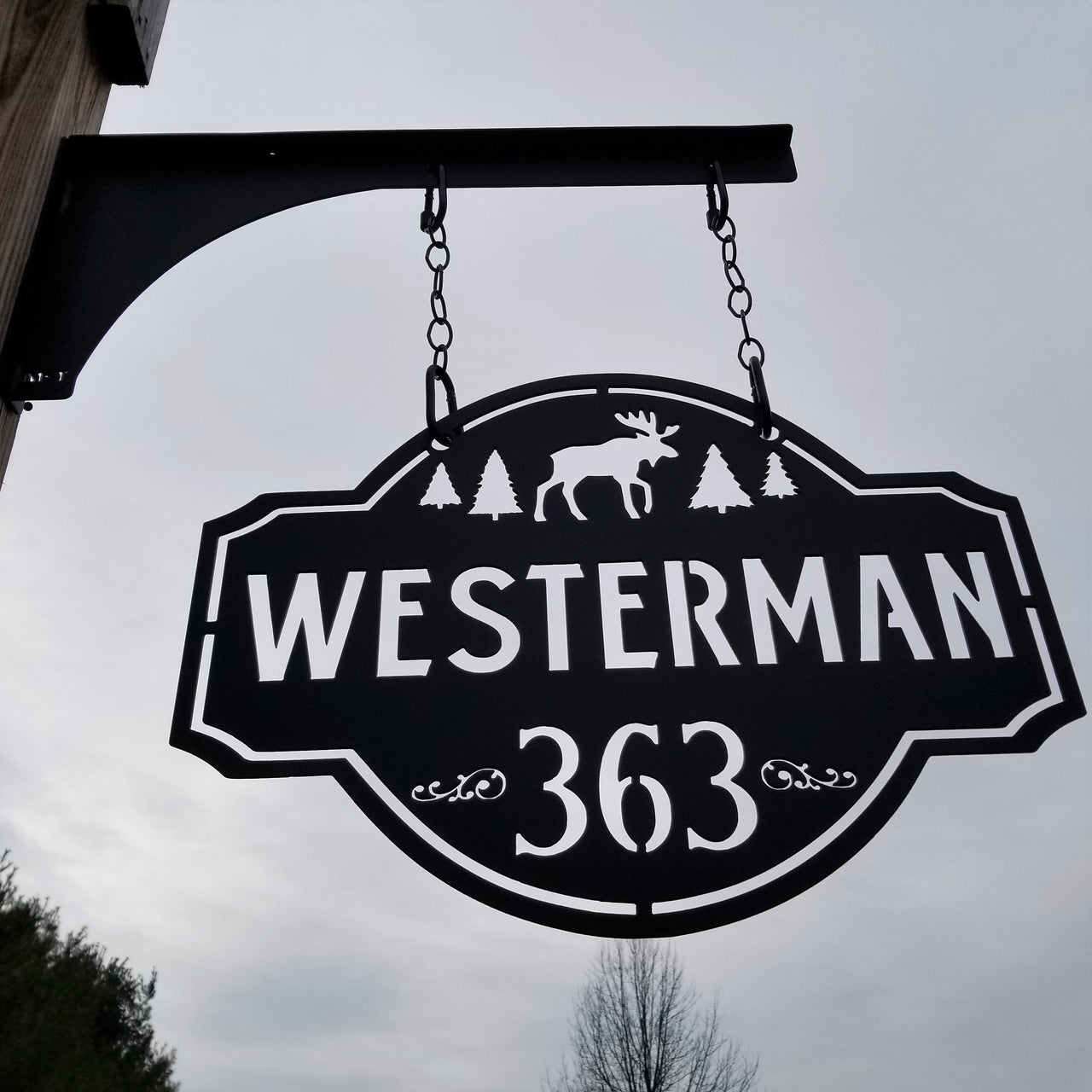 This is a personalized metal address sign that hangs from a hanging bracket mounted to a wooden post.  The sign is powder coated black and features a forest scene with a moose in the center. The sign reads, "Westerman 363".