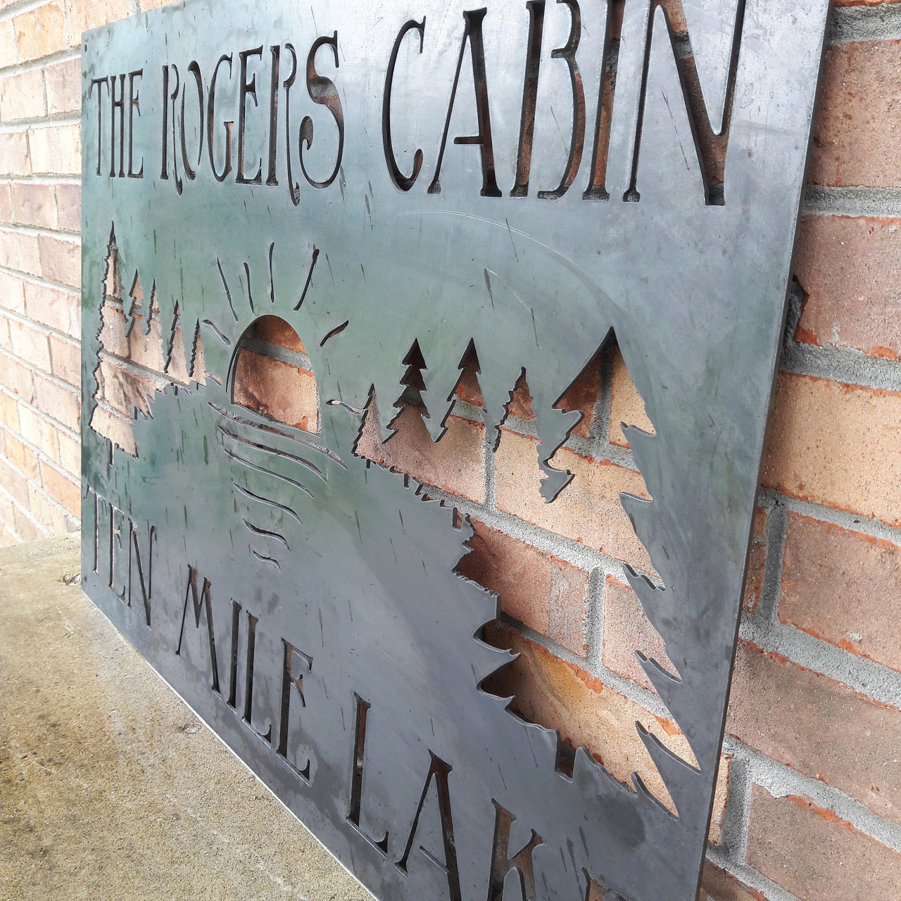 This metal sign has a tree line with a setting sun and reads, "The Rogers Cabin Ten Mile Lake"