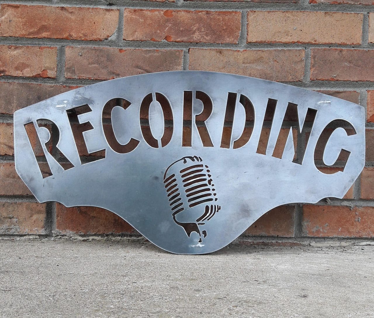 This metal sign has hidden mounts welded to the back which stands the sign 1" off the wall.  There is an image of a microphone on the sign and a line of text. The sign reads, "RECORDING".
