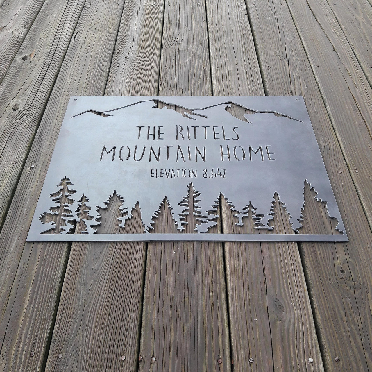 rectangular sign with a mountain range across the top and a coniferous tree line. There are two lines of text on this sign as well as an elevation.  The sign reads, " The Rittels Mountain Home, Elevation 8,647" 