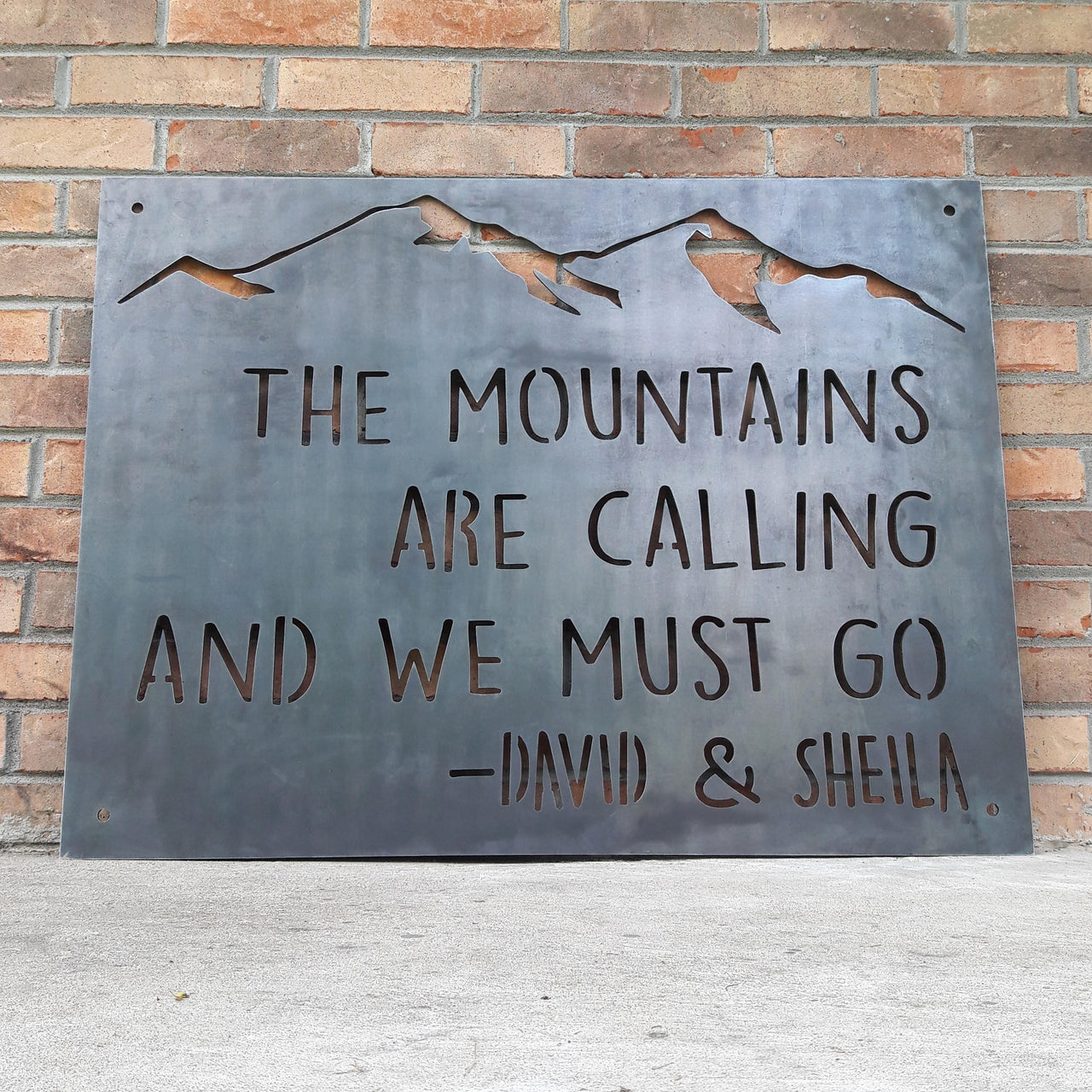 This Metal sign has a mountain range across the top and features a quote by John Muir. The sign reads, "The Mountains Are Calling And I must Go, David and Sheila".