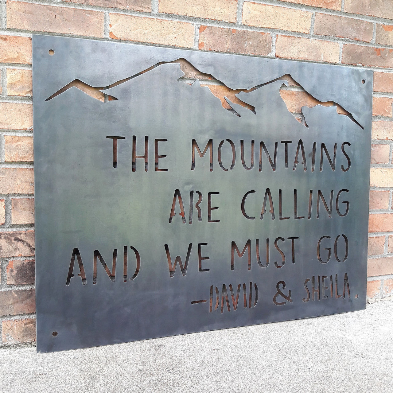 This Metal sign has a mountain range across the top and features a quote by John Muir. The sign reads, "The Mountains Are Calling And I must Go, David and Sheila".