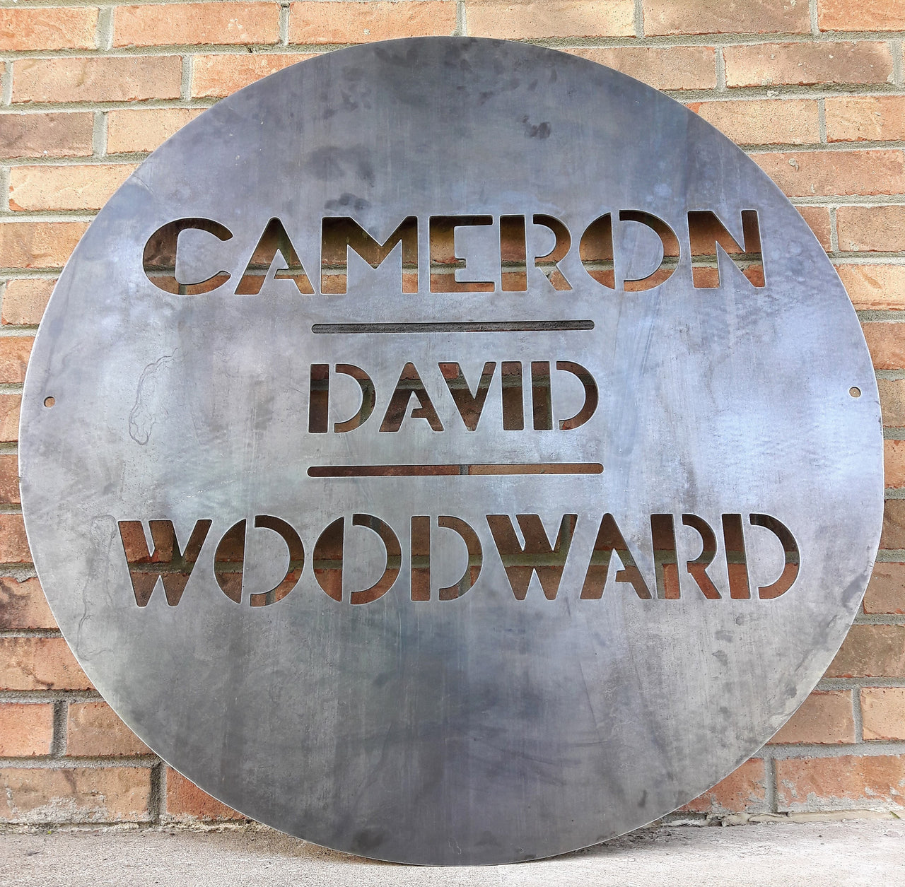 This is a round art deco sign that has three lines of text with a straight line seperating them. The sign reads, "Cameron David Woodward"