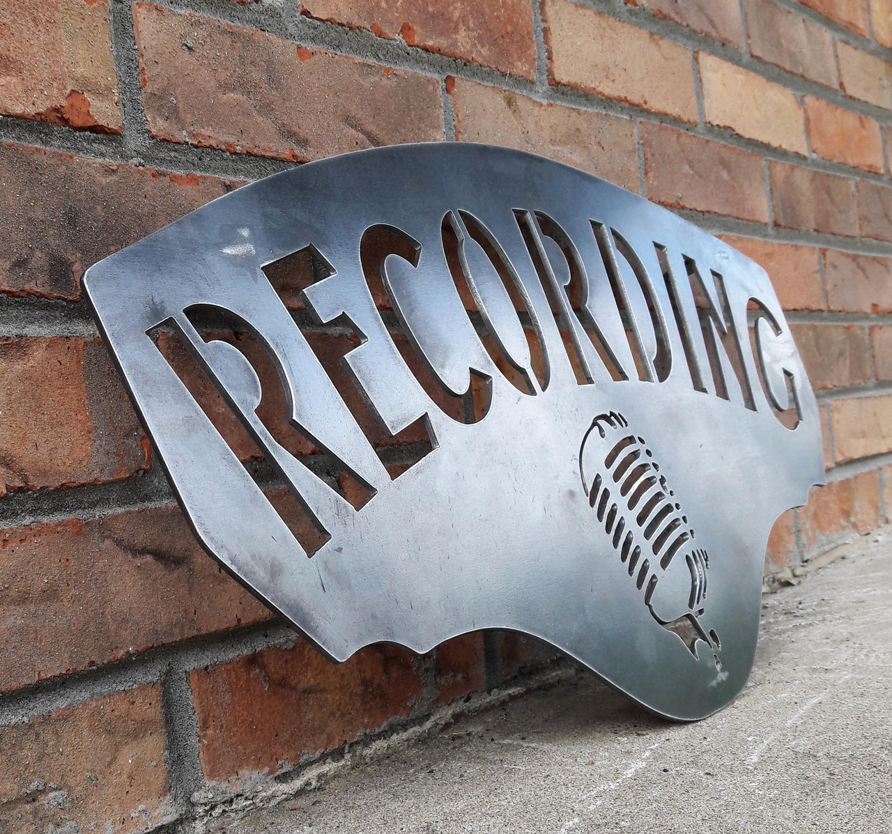 This metal sign has hidden mounts welded to the back which stands the sign 1" off the wall.  There is an image of a microphone on the sign and a line of text. The sign reads, "RECORDING".