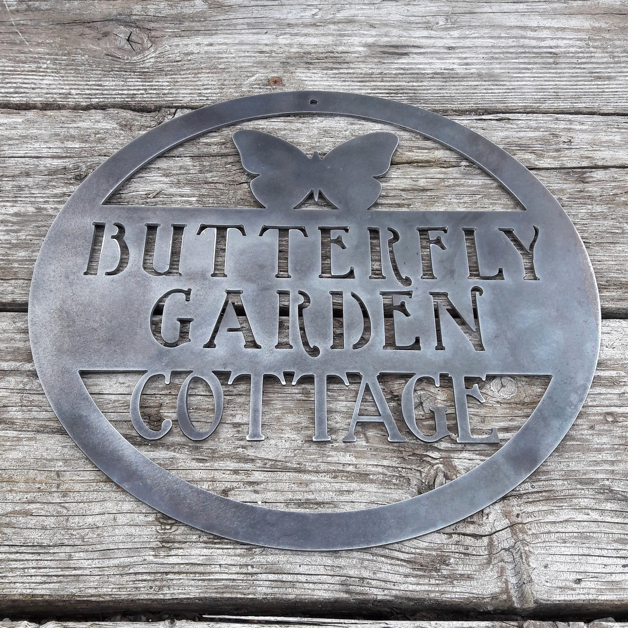 Round metal sign with the image of a Butterfly at the top and three lines of text. The sign reads, " Butterfly Garden Cottage".