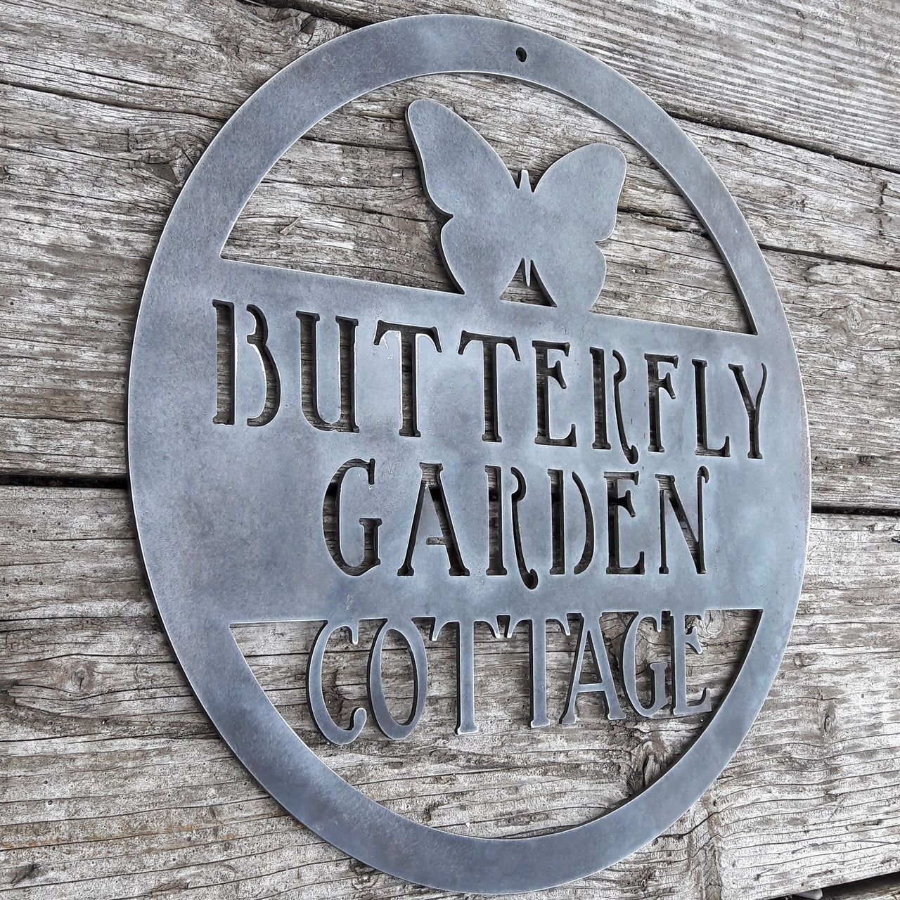 Round metal sign with the image of a Butterfly at the top and three lines of text. The sign reads, " Butterfly Garden Cottage".